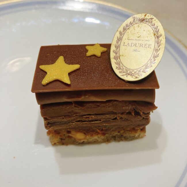 ラデュレ日比谷『星の王子さま』コラボアフタヌーンティーのミニパティスリー「プレジール・シュクレ」。四角いミルクチョコレートが層になっており、上部には星型の飾りとラデュレのオーバル型のロゴプレートが添えられている。土台はヘーゼルナッツを使用している。