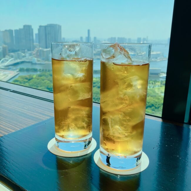 コンラッド東京のバー＆ラウンジで提供されている、コンラッド東京オリジナルアイスティー。グラスの背景には東京湾と都会のビル群の絶景が広がり、テーブルには氷が入ったグラス2つが並べられ、グラスの縁には白いコースターが敷かれている。