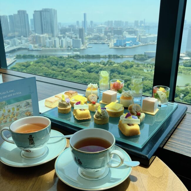コンラッド東京 バー＆ラウンジ トゥエンティエイトのアフタヌーンティー。大きな窓の外には東京湾や浜離宮恩賜庭園を見下ろす絶景が広がり、手前のガラスプレートにはウェッジウッドの食器とともに彩り豊かなスイーツとセイボリーが並んでいる。