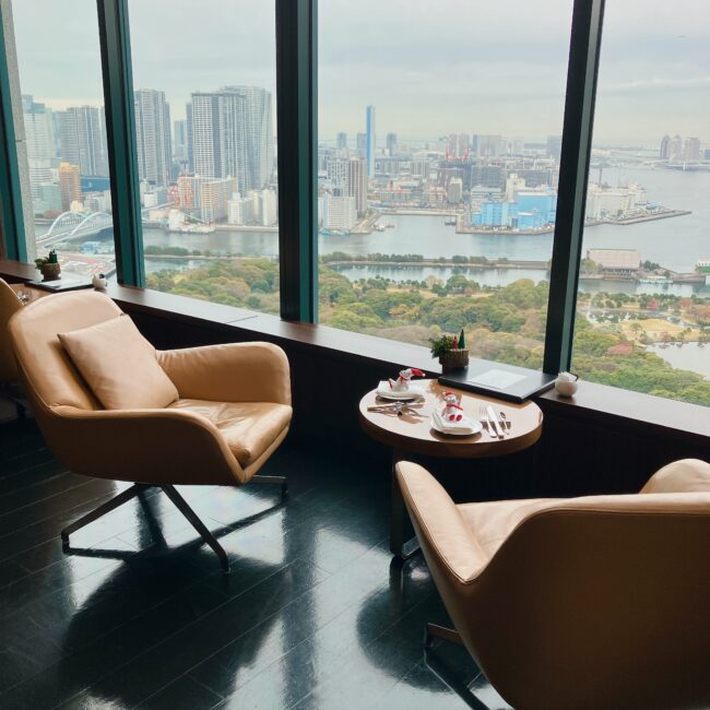 コンラッド東京 バー＆ラウンジ トゥエンティエイトの窓際席。革張りのベージュ色のハイバックチェアが2脚、丸いテーブルを挟んで向かい合って配置されており、大きな窓からは浜離宮恩賜庭園と東京湾、都会のビル群が見下ろせる。
