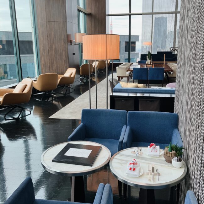 コンラッド東京 バー＆ラウンジ トゥエンティエイトの店内。深い青色のチェアと大理石のテーブルが手前にあり、奥には木目調の壁と大きなフロアランプが配置されている。全体的にダークトーンの床と窓からの光で、都会的で洗練された雰囲気が漂っている。