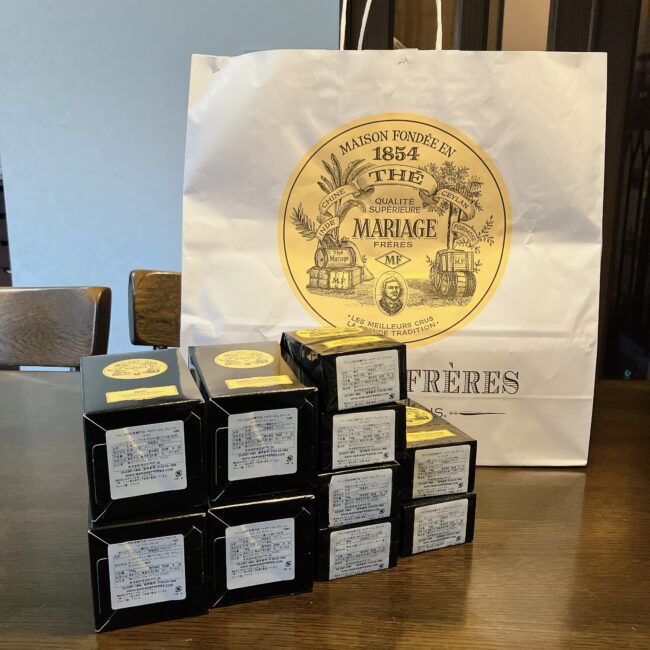 マリアージュフレール(マリフレ)の10銘柄福袋の中身の写真。ティーバッグ(または小型缶)の紅茶が10個積み上げられており、背景にはマリフレの紙袋が見える。福袋の開封レビュー。