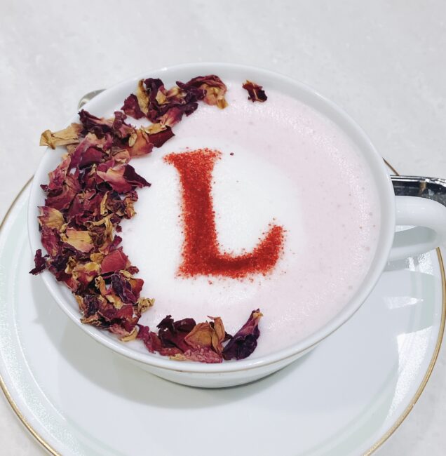 ラデュレのイスパハンのラテ。ピンク色のラテアートに赤いパウダーで「L」の文字が描かれ、カップの縁には多数のドライローズの花びらが散らされている。