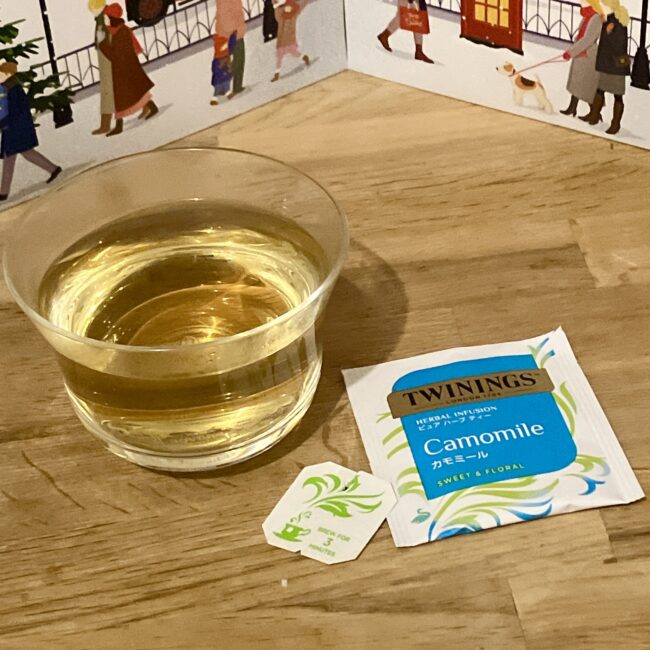 トワイニング アドヴェントカレンダー2025の2日目、カモミールのティーバッグと、ガラスのカップに入った黄金色の水色（ハーブティー）。リラックスタイムに最適なカモミールティー。ティーバッグには「3 MINUTES」の文字が見える。