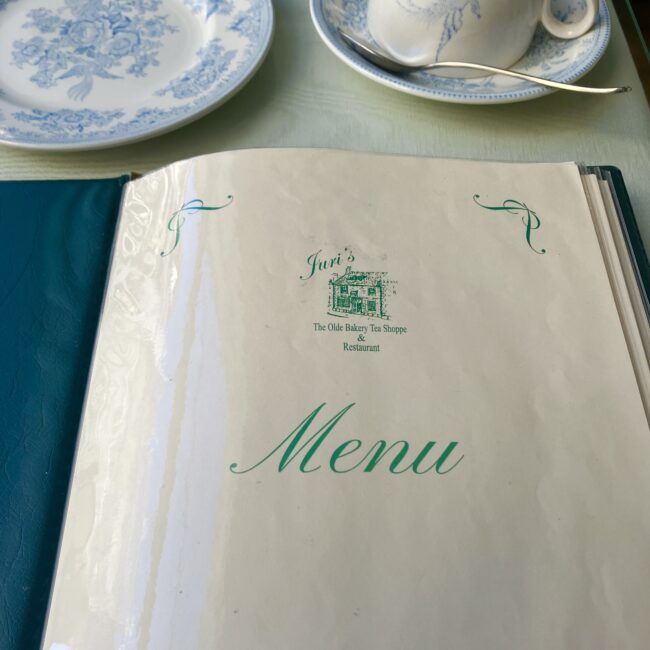 ジュリスティールームのメニュー表紙。表紙中央には、ウィンチカム時代の店舗のイラストと「Juri's The Olde Bakery Tea Shoppe & Restaurant」のロゴ。ブルー&ホワイトの皿とカップが写っている。