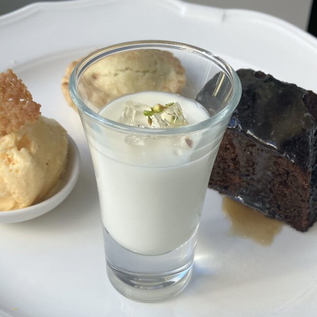 ジュリスティールームの生クリームとエルダーフラワーのポセット。小さなグラスに入った白いデザートで、奥にはミンスパイとステッキートフィープディングが見えてい