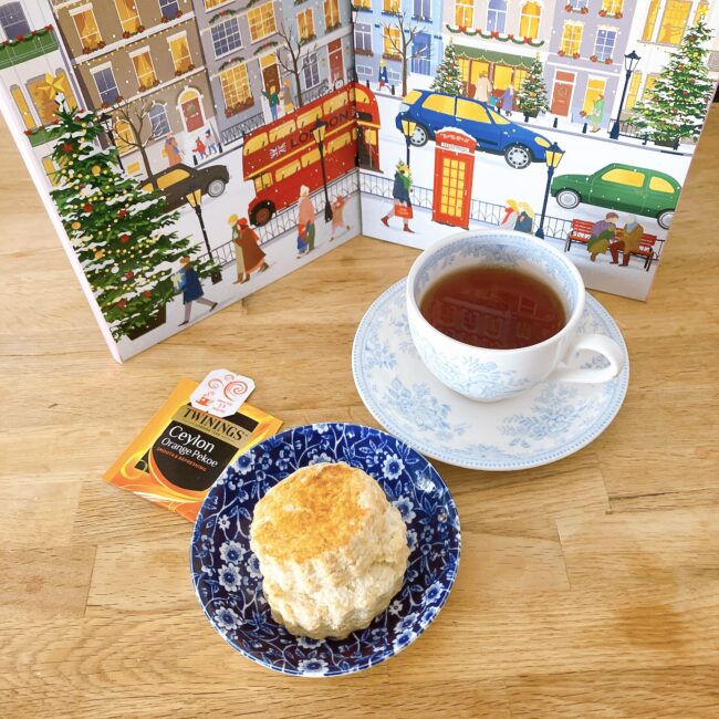 ロンドンの冬の街並みが描かれたトワイニングのアドヴェントカレンダーの前に、繊細なブルーの模様が入ったティーカップと、濃紺の柄皿にのった焼きたてのスコーンが並ぶ。手前にはオレンジ色のトワイニング「セイロン オレンジ ペコ」の個包装が置かれている。