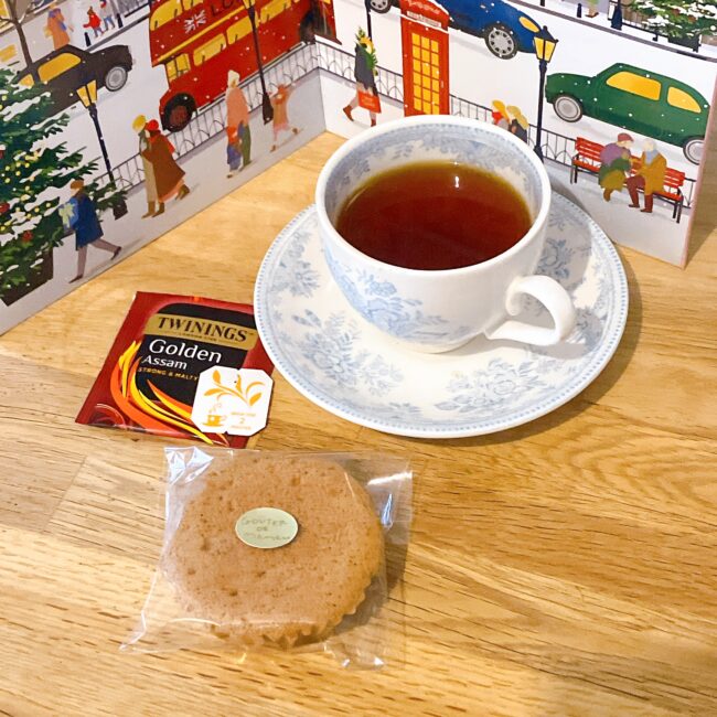 ロンドンの冬の街並みが描かれたトワイニングのアドヴェントカレンダーを背に、上品なブルーの花柄ティーカップに注がれた「ゴールデン アッサム」。手前には透明な袋に入った紅茶マドレーヌと、トワイニングの赤い「Golden Assam」の個包装が添えられている。