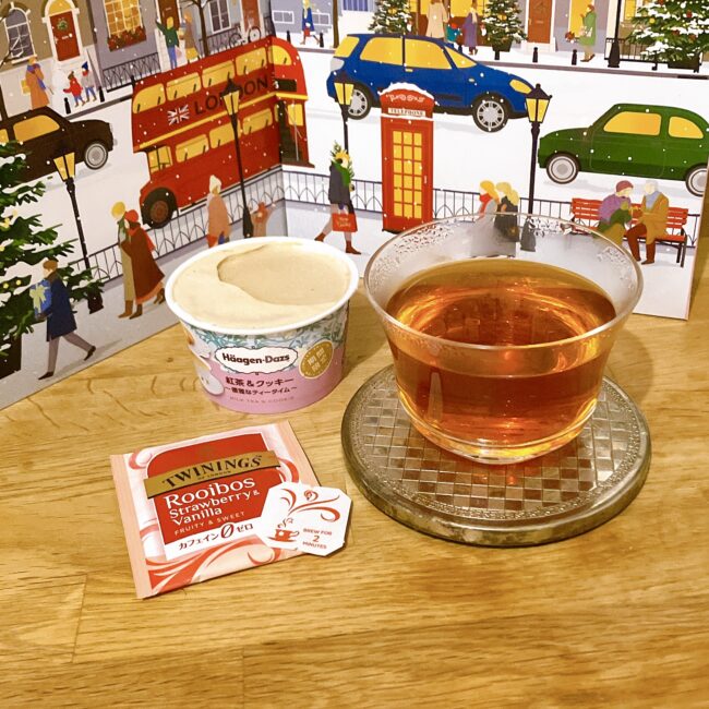 ロンドンの冬が描かれたアドヴェントカレンダーを背景に、耐熱ガラスのカップに注がれた輝くルイボスティー。隣にはハーゲンダッツの期間限定「紅茶＆クッキー」のアイスクリーム、手前にはトワイニングの赤い「Rooibos Strawberry & Vanilla」の個包装が置かれている。