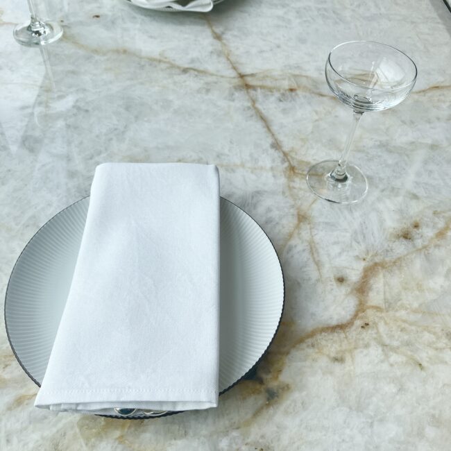 白い大理石のテーブルの上に置かれた、真っ白なプレートとナプキン。ナプキンはカトラリーを包み込むように丁寧に折られており、隣には繊細なシャンパングラスが添えられている。