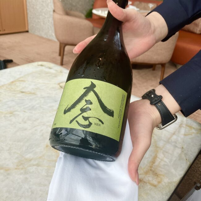 スタッフが「念(NEN)」というラベルの付いた玄米茶スパークリングティーのボトルをサーブしている様子。