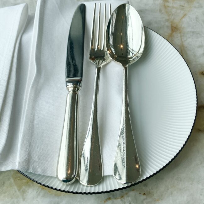 ナプキンから現れたシルバーのカトラリー(ナイフ、フォーク、スプーン)。光沢のある美しい銀食器が、縁に細かい筋模様が入った上品な白いプレートの上に並んでいる。