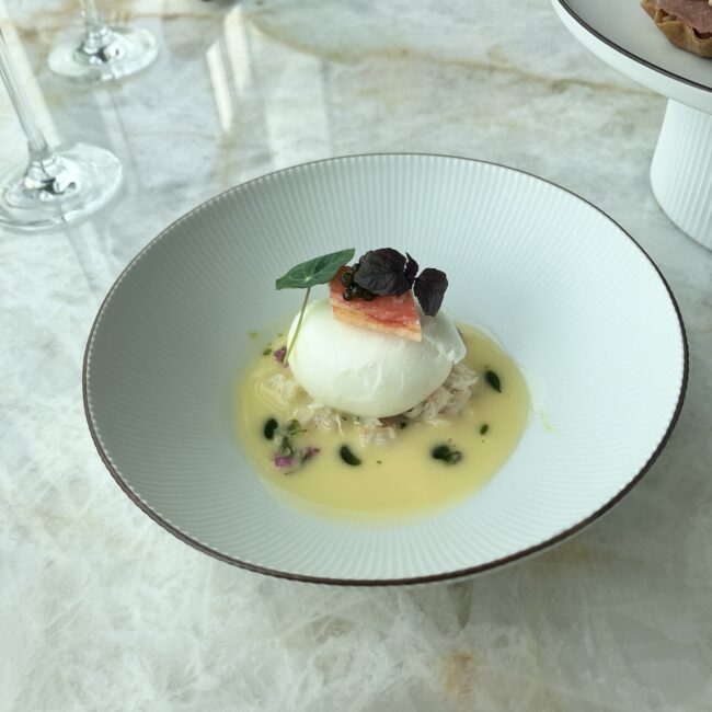 白い丸皿に盛り付けられたズワイ蟹のポーチドエッグ。上にキャビアとハーブが添えられ、周囲には黄色いブールブランソースが広がっている様子。