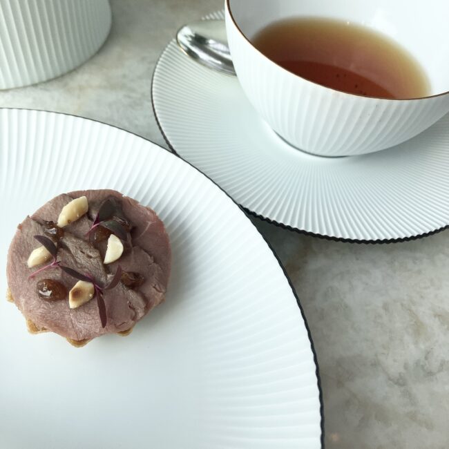 鴨胸肉のスモークとイチジクのタルトを乗せた白い皿と、その奥に並ぶ琥珀色のドアーズ(紅茶)。タルトの上にはナッツとカシスの実が散らされ、洗練されたペアリングの光景。