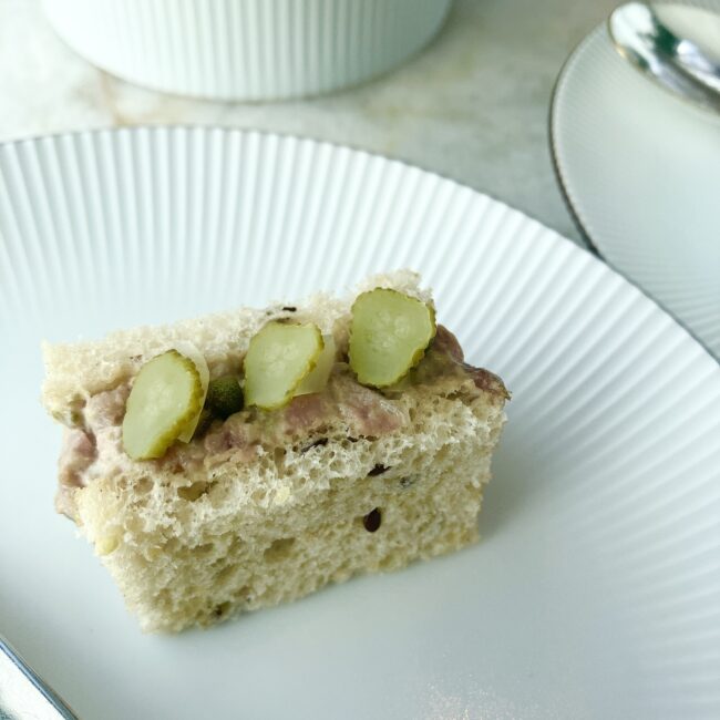 ライブレッドに挟まれたローストビーフのサンドイッチ。断面にはケッパーやオニオンが見え、上には3つの輪切りピクルスが等間隔に並んでいる。