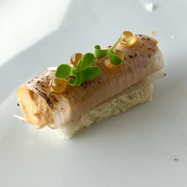 白いパンの上に乗せられた下仁田葱のグリル。円筒状に巻かれた葱の中にアーモンドソースが詰まっており、上にはオレンジ色のオイルの粒と若芽が添えられている。