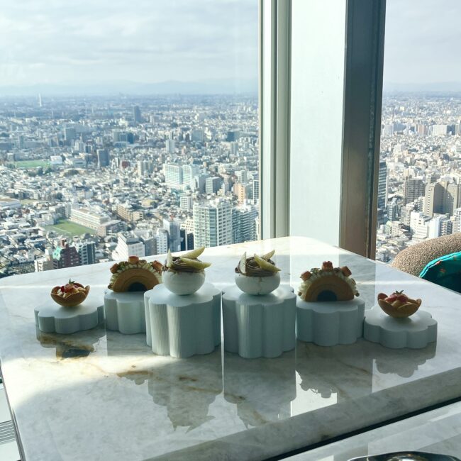 西新宿のパノラマビューを背景に、白い陶器のスタンドに立体的に配置されたアフタヌーンティーの全景。モダンで建築的なプレゼンテーション。
