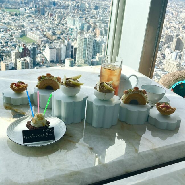 窓外に広がる東京の広大なパノラマを背景に、テーブルいっぱいに並んだアフタヌーンティーのスタンドと「ハッピーバースデー」のメッセージプレート。祝福のムード溢れる華やかな光景。