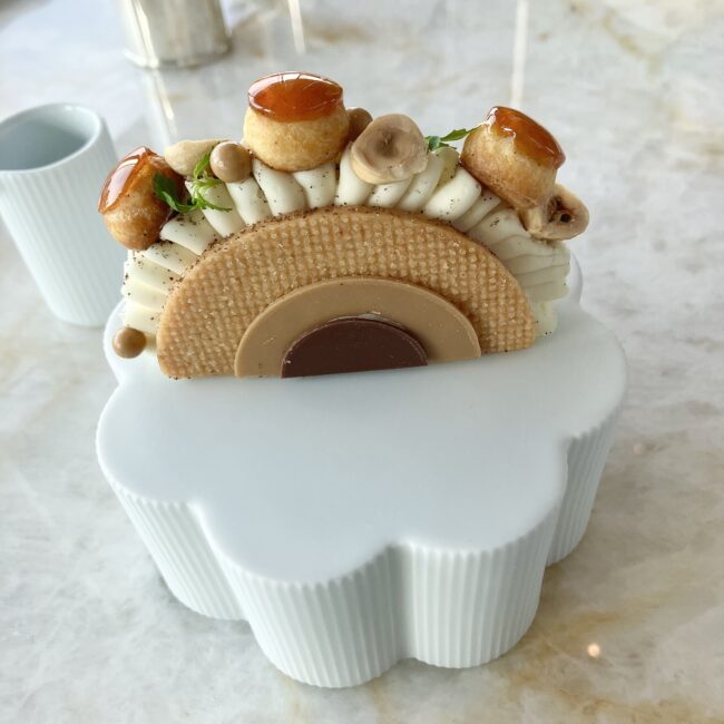 橋のようなモダンな構造のサントノーレ。波打つように絞られたバニラクリームの上に、キャラメリゼされて艶やかに輝くプチシューとヘーゼルナッツが整然と並ぶ。側面は半円形のビスケットとチョコレートプレートで装飾されている。