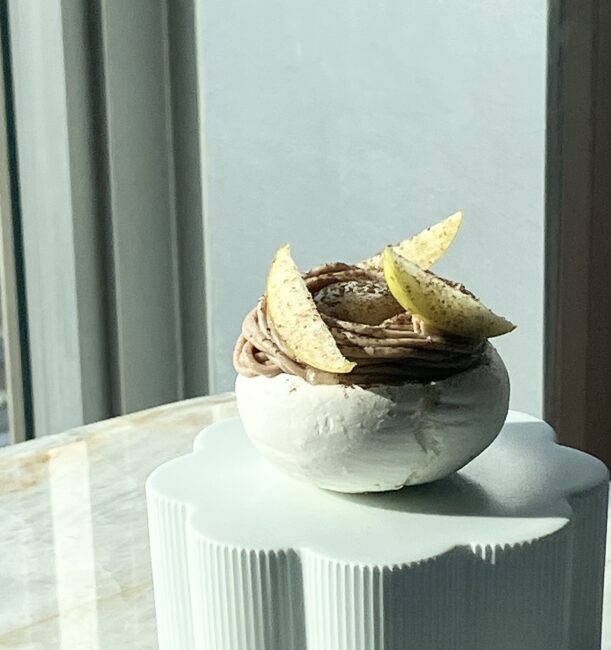 白いメレンゲを器に見立てた、建築的な造形の和栗のモンブラン。渦巻状に絞られたマロンクリームの上に、薄くスライスされた梨とスパイスが飾られている。窓からの自然光が、メレンゲの質感と立体的な構造を際立たせている。