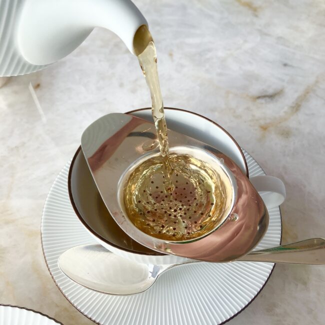 パークハイアット東京「ピークラウンジ」の紅茶提供スタイル。白いティーポットから、銀色のティーストレイナー(茶こし)を通して、琥珀色の紅茶がカップに注がれている様子。