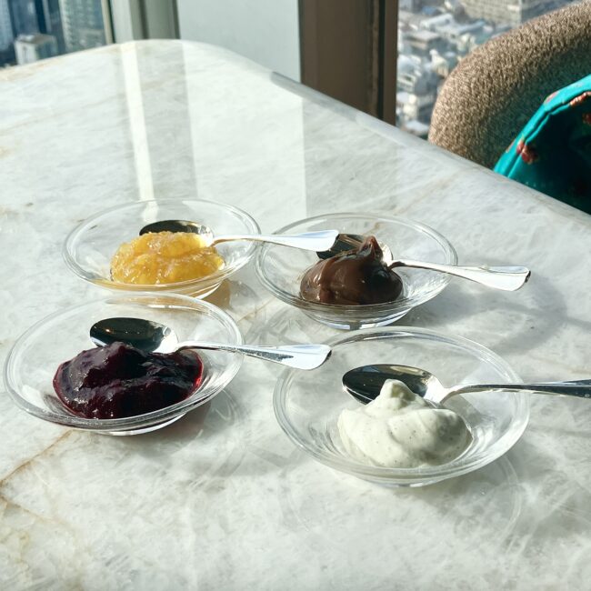パークハイアット東京「ピークラウンジ」で提供される4種類のコンディメント。光が差し込む大理石のテーブルに、マーマレード、いちごジャム、クリームチーズ、チョコレートクリームがそれぞれ小さなガラスの器に入れられ、スプーンが添えて並べられている。