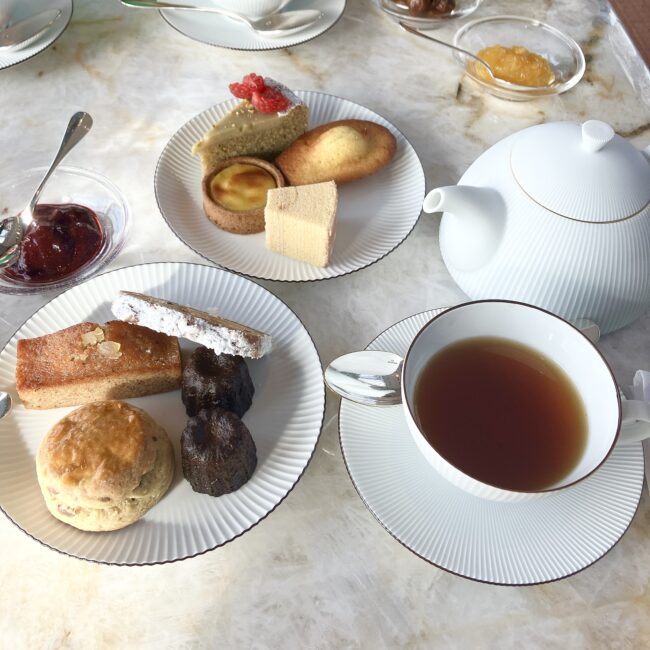ワゴンから選んだ9種類の焼き菓子を「全部載せ」した、贅沢なティータイムのテーブル風景。白い大理石のテーブルに、2皿に盛り分けられたスイーツ、白い磁器のティーポット、ティーカップが並んでいる。