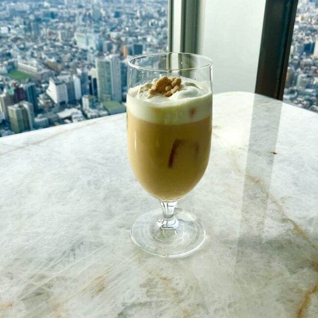 ピークラウンジの新メニュー「アイスロイヤルミルクティー」。濃厚なミルクティーの上にふんわりとしたフォームとクランブルが乗り、背景には東京のビル群が広がる。