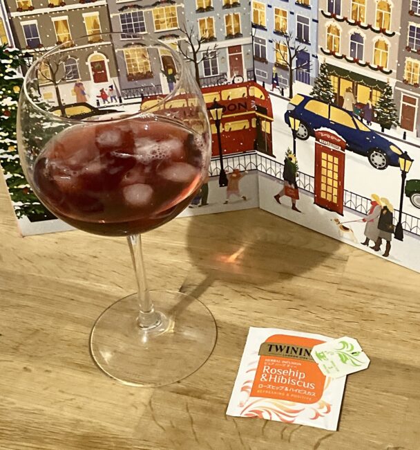 木目のテーブルに置かれた大きなワイングラス。中には氷がたっぷり入った鮮やかな赤色のハーブティーが注がれている。手前にはトワイニングの「ローズヒップ＆ハイビスカス」のティーバッグの個包装があり、背景にはイギリスの街並みが描かれたアドヴェントカレンダーが広げられている。