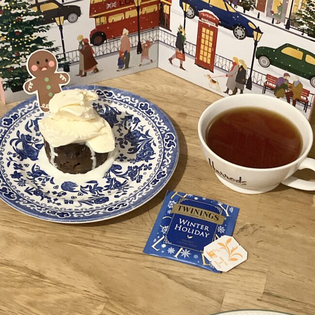 木製のテーブルの上に置かれた、クリスマスイブのティータイムの風景。トワイニングの「ウィンターホリデー」のティーバッグと、Harrods（ハロッズ）のロゴが入ったカップに注がれた深い琥珀色の紅茶。隣には、白いアイスクリームがたっぷり載ったクリスマスプディングの皿が添えられている。背景には、ロンドンの街並みや赤い2階建てバスが描かれた華やかなアドヴェントカレンダーが広がり、温かく賑やかなホリデーの雰囲気を演出している。