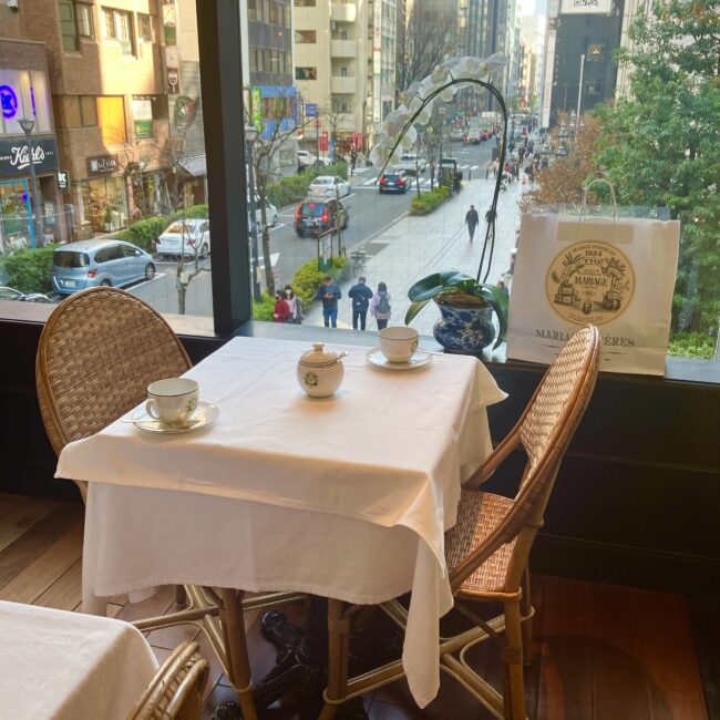 マリアージュフレール銀座松屋通り店のティーサロン。大きな窓から銀座の街並みを見渡せる開放的な窓際席。テーブルには白いクロスとティーカップ、傍らにはマリアージュフレールの福袋が置かれ、おひとりさまランチの贅沢な時間が流れる様子。