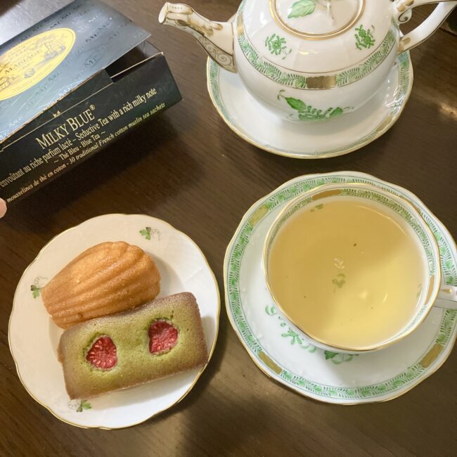 ヘレンドの「ウィーンの薔薇」のティーセットに注がれたミルキー ブルー。隣の小皿には、アールグレイのマドレーヌと抹茶とラズベリーのフィナンシェが添えられている。左端にはミルキー ブルーの黒い箱が見える。