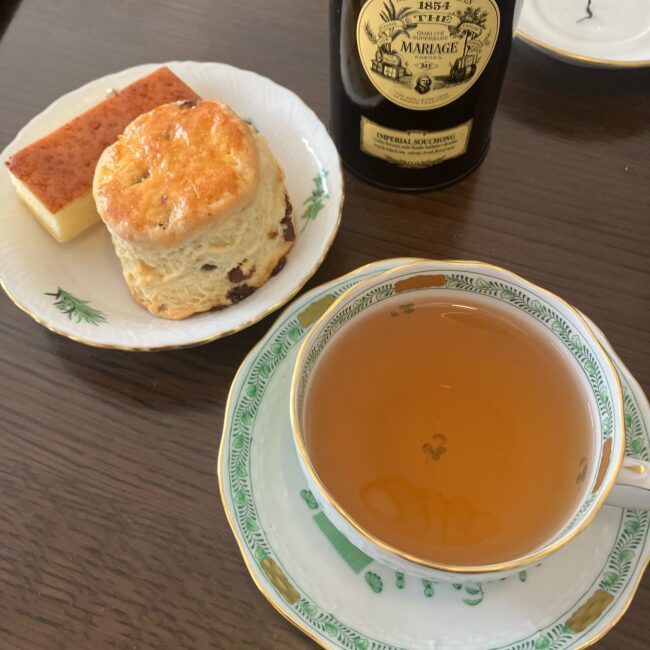 マリアージュ フレールの紅茶缶「インペリアル スーチョン」と、ティーカップに注がれた明るい琥珀色の紅茶。隣の皿には、ふっくらと焼き上がったレーズンスコーンと四角い焼きチーズケーキが添えられている。