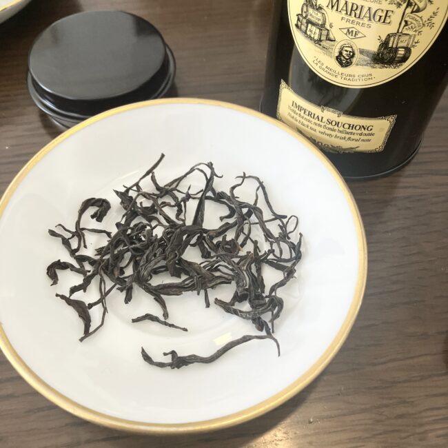 白い小皿に広げられた、マリアージュ フレール「インペリアル スーチョン」の茶葉。黒く艶があり、細長く美しくよじれた大ぶりの茶葉が特徴的。背景には黒い紅茶缶が配置されている。
