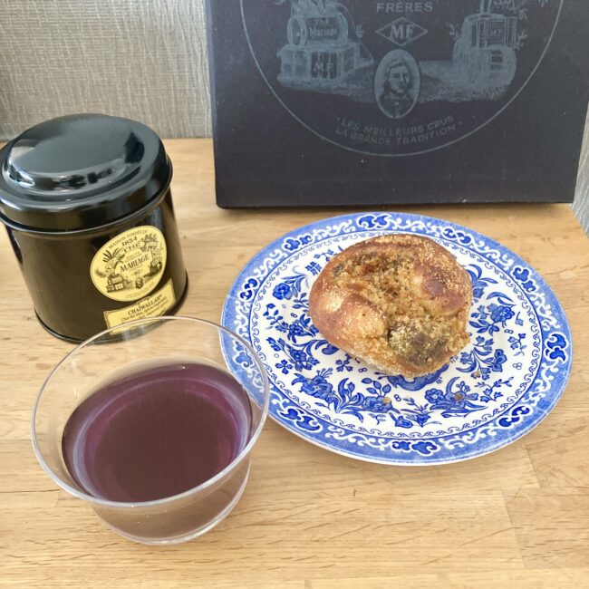 マリアージュフレールの紅茶缶「チャイワラ (CHAIWALLAH)」と、抽出された鮮やかな紫色のチャイ。手前にはペアリングされたカレーパンが青い皿に乗せられており、スパイスの香りが漂ってきそうなティータイムの様子。