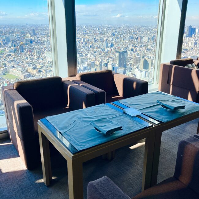 2023年改修前のパークハイアット東京・ピークラウンジ。窓側の席には青いテーブルクロスがかけられ、重厚なブラウンのソファが並んでいる。テーブルの上にはナプキンとカトラリーがセットされ、窓の外には冬の澄んだ青空と新宿の街並みが広がっている。