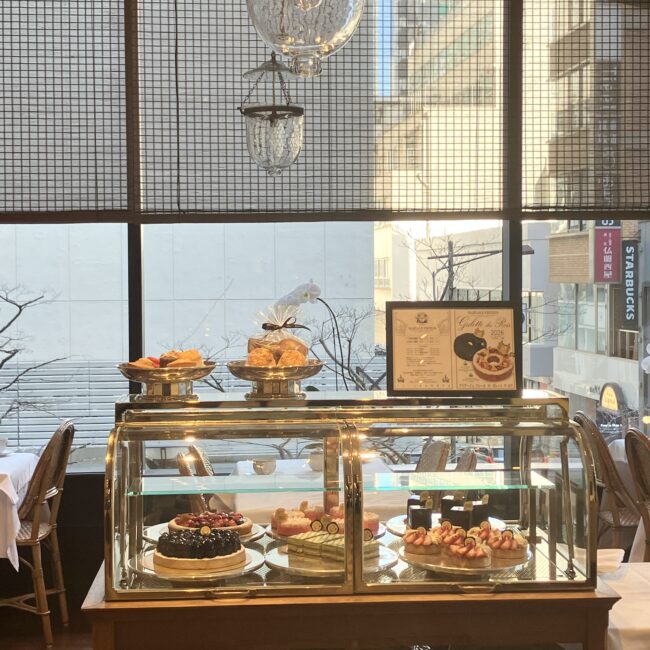 マリアージュフレール銀座松屋通り店のスイーツワゴン。ショーケースにはお茶を使用したケーキな並び、ワゴンの上にはテイクアウト用の焼き菓子セットや期間限定ガレットデロワのポスターが飾られている。