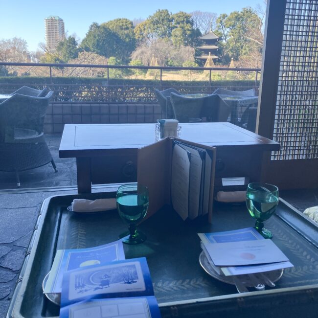 ル・ジャルダンの窓際席からの眺望。テーブルには光を反射するグリーンのグラスとメニューが並び、その向こうには冬の青空に映える椿山荘の三重塔と広大な庭園が広がっている。