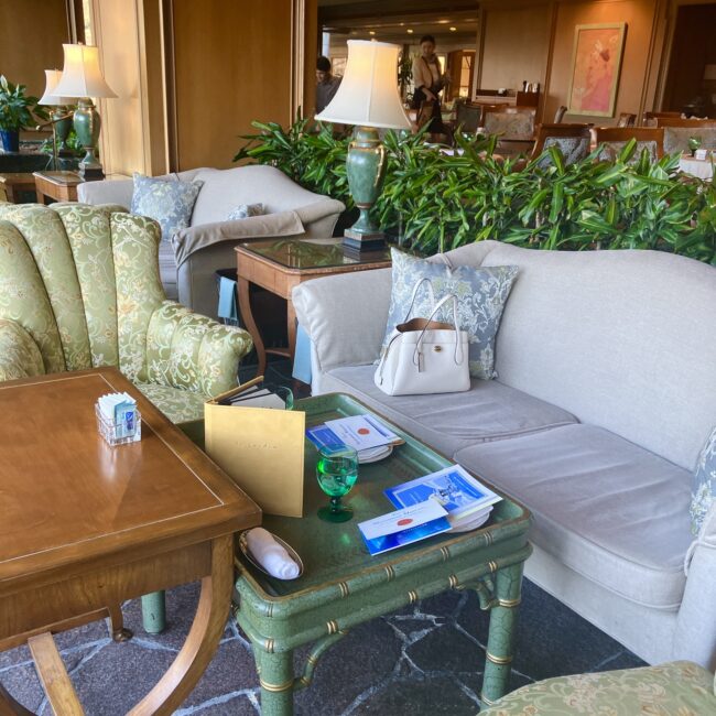 窓際のソファ席にセッティングされたアフタヌーンティーのテーブル全景。グリーンのトレイ、専用メニュー、白いバッグを置いても余裕のある広々としたパーソナルスペースが確認できる。