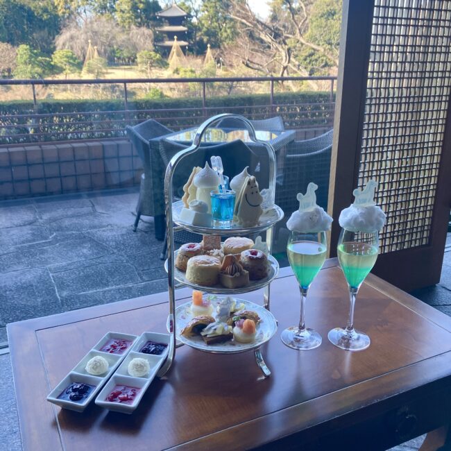 椿山荘「ル・ジャルダン」の窓際席に用意された、ムーミンアフタヌーンティーの3段スタンド。背景には冬の庭園と三重塔が広がり、手前にはオーロラ色のウェルカムドリンクと綿あめを添えたグラスが並ぶ、優雅なティータイムの全景。