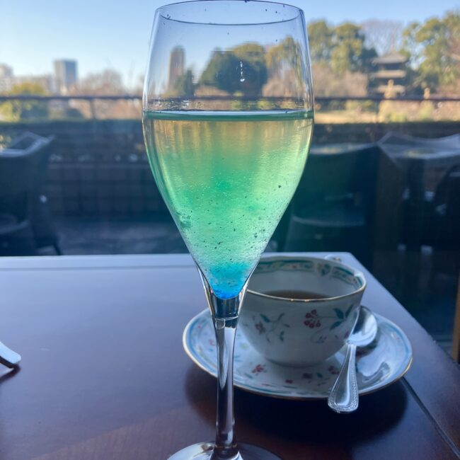 椿山荘ル・ジャルダンの「オーロラ」をイメージしたウェルカムドリンク。青から黄色への美しいグラデーションが映えるグラスの背後に、ロンネフェルトの紅茶が注がれた上品なティーカップが置かれている。背景には冬の庭園が広がる。冬の静寂の中に春を待つ生命の息吹を感じさせる一瞬