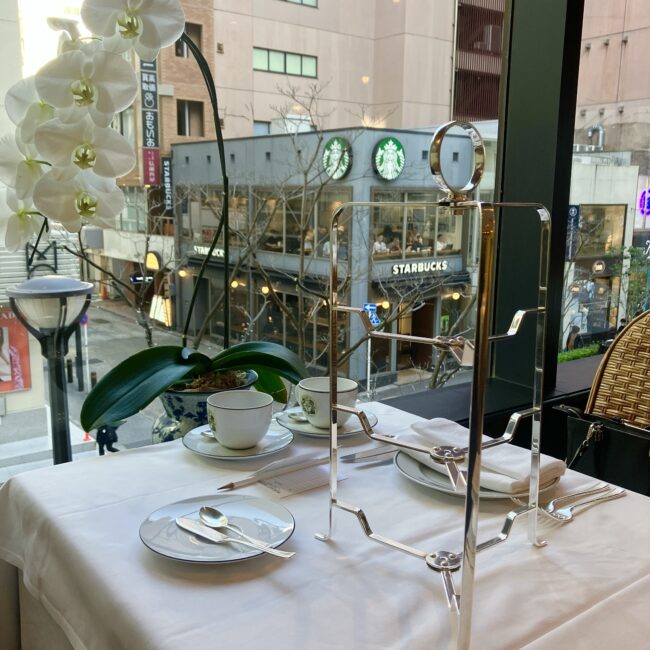 マリアージュフレール銀座松屋通り店2階の窓際席。シルバーの3段スタンドと白い胡蝶蘭が置かれ、大きな窓の外には銀座の街並みとスターバックスが見える開放的な空間。