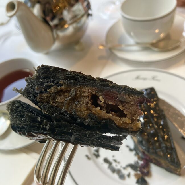 フォークで持ち上げられた黒いガレット・デ・ロワの断面。幾重にも重なる薄い黒いパイ生地の中に、しっとりとした茶色のアーモンドクリームが詰まっており、アクセントの赤いベリーが見える。