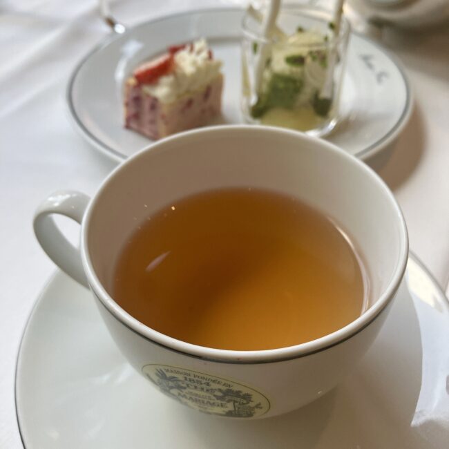 マリアージュフレールのベトナム産緑茶「モンダンドシヌ (MONTAGNE D'INDOCHINE) Ref. T4909」。白磁のカップに注がれた水色は、透明感のある淡い黄金色。奥にはペアリングされたスイーツ（ヴェリーヌとビュッシュ）が少し写り込んでいる。