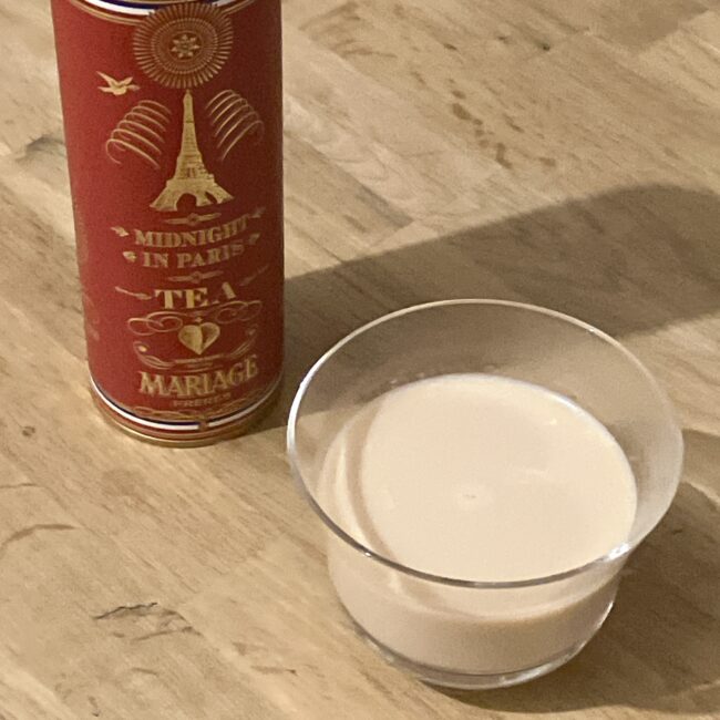 透明なガラスカップにたっぷりと注がれた「ミッドナイト イン パリ」のミルクティー。温かみのあるクリーミーなベージュ色をしており、濃厚な質感が伝わる。背景には金色のエッフェル塔が描かれたマリアージュフレールの赤い紅茶缶が置かれている。