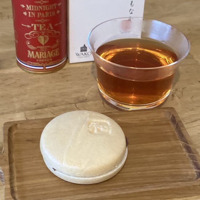 木製のコースターに乗った白くて丸い和光の最中と、ガラスのカップに注がれた赤褐色のマリアージュフレールのルイボスティー「ミッドナイト イン パリ」。背景には赤い紅茶缶と和光の白い箱が置かれている。