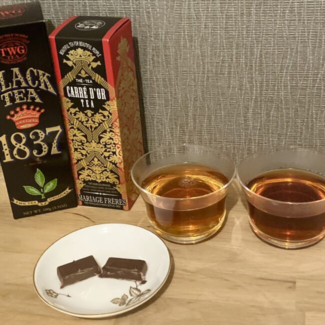 イナムラショウゾウのショコラと紅茶のペアリング検証風景。TWG Tea「1837 BLACK TEA」とマリアージュフレール「カレ ドール」のパッケージ、グラスに注がれた紅茶、手前の皿に並ぶバニラとベルガモットのショコラ