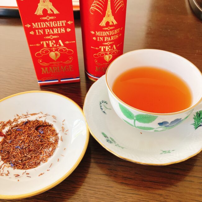 花柄のティーカップに注がれた美しいオレンジ色の「ミッドナイト イン パリ」のストレートティー。手前の小皿にはルイボスの茶葉が出されており、赤茶色の茶葉の中に青い矢車菊の花びらが混ざっている様子が確認できる。背景には赤い外箱と紅茶缶が並んでいる。