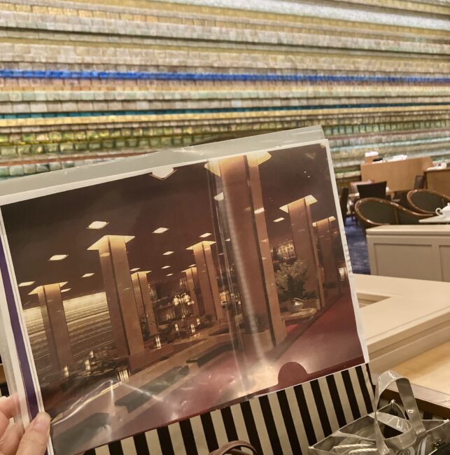 帝国ホテル100年史の資料（平成の大改修時の写真）を、現在のランデブーラウンジの背景とかざして比較している様子。