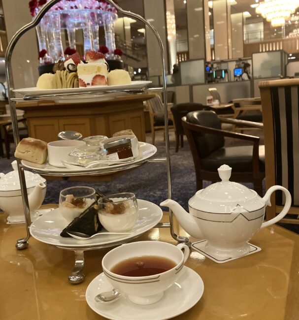 帝国ホテル 東京「ランデブーラウンジ」のアフタヌーンティー。3段スタンドの上段には苺のショートケーキやモンブラン、中段にスコーンとジャム、下段にセイボリーが並ぶ。手前には白いティーポットと紅茶が入ったカップ、背景には特徴的な「光の壁」とシャンデリア「ゴールデンローズ」が見えるテーブルの様子。